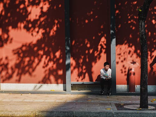 Candid Photo of a Boy Sitting on a Bench Outside of a Red Building with a Shadow of a Tree