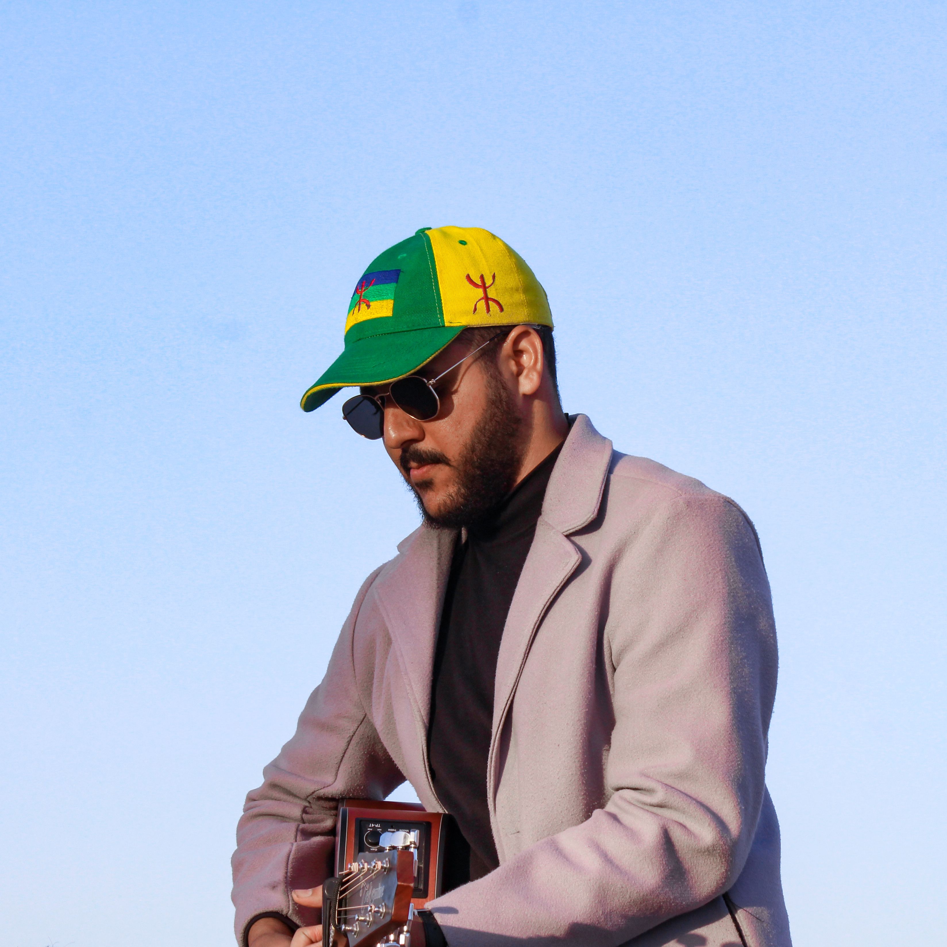 A stylish man wearing sunglasses and a colorful cap playing guitar outdoors.