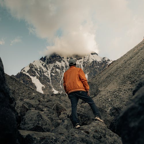 Back View of a Person in Orange Jacket Standing on Rocky Mountain under the Cloudy Sky