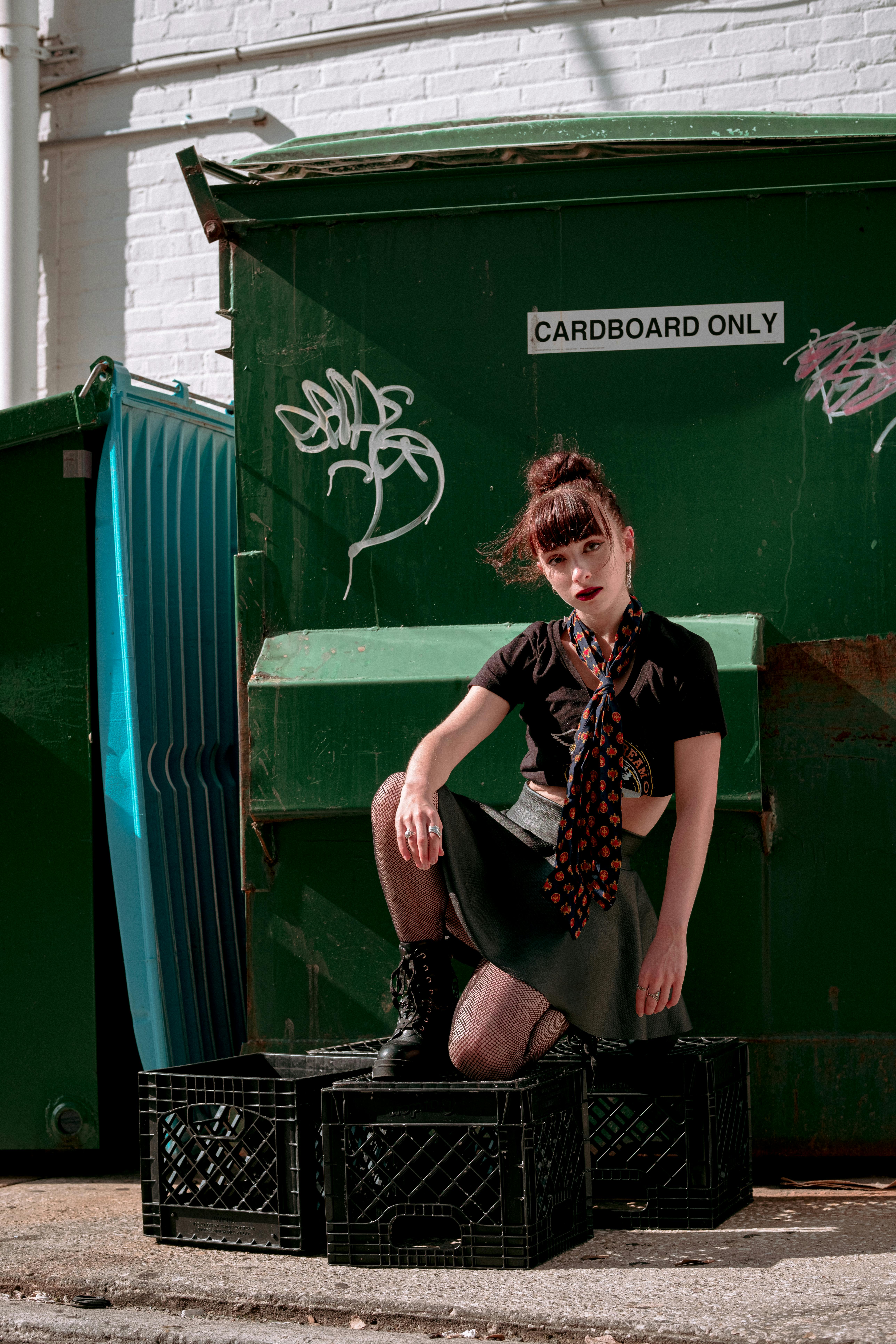 a woman sitting on a crate in front of a green trash can