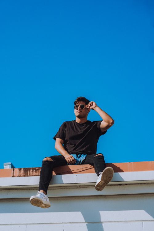 Free Young man in sunglasses sitting on rooftop edge under clear blue sky in Orlando. Stock Photo