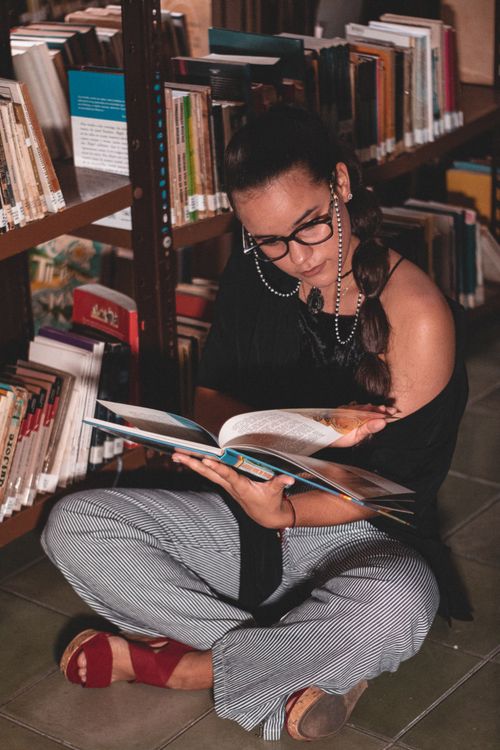 Free Woman seated on library floor, deeply engrossed in a book, surrounded by bookshelves. Stock Photo