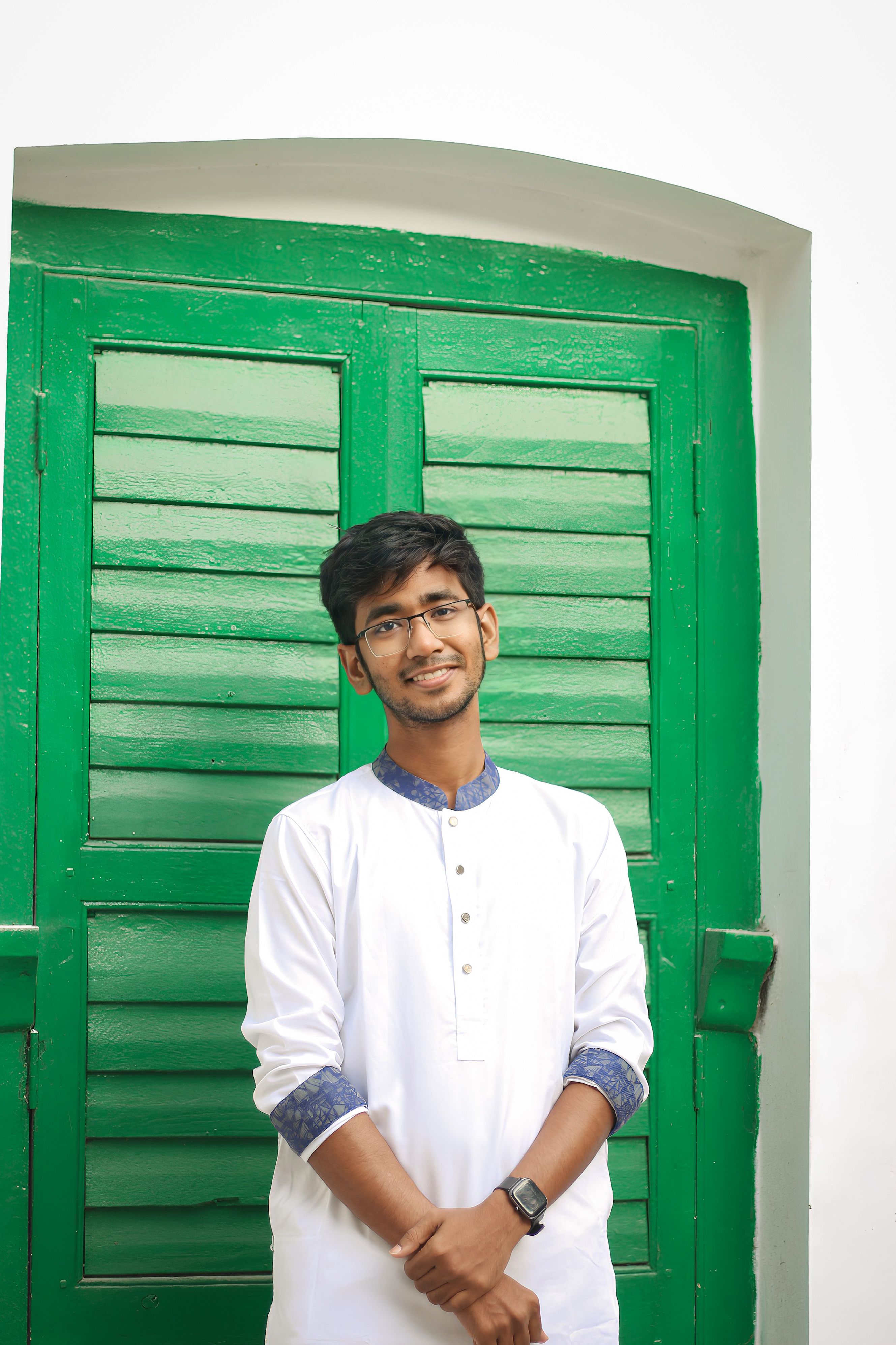 Portrait of a young man in a white kurta smiling by a green shuttered window.