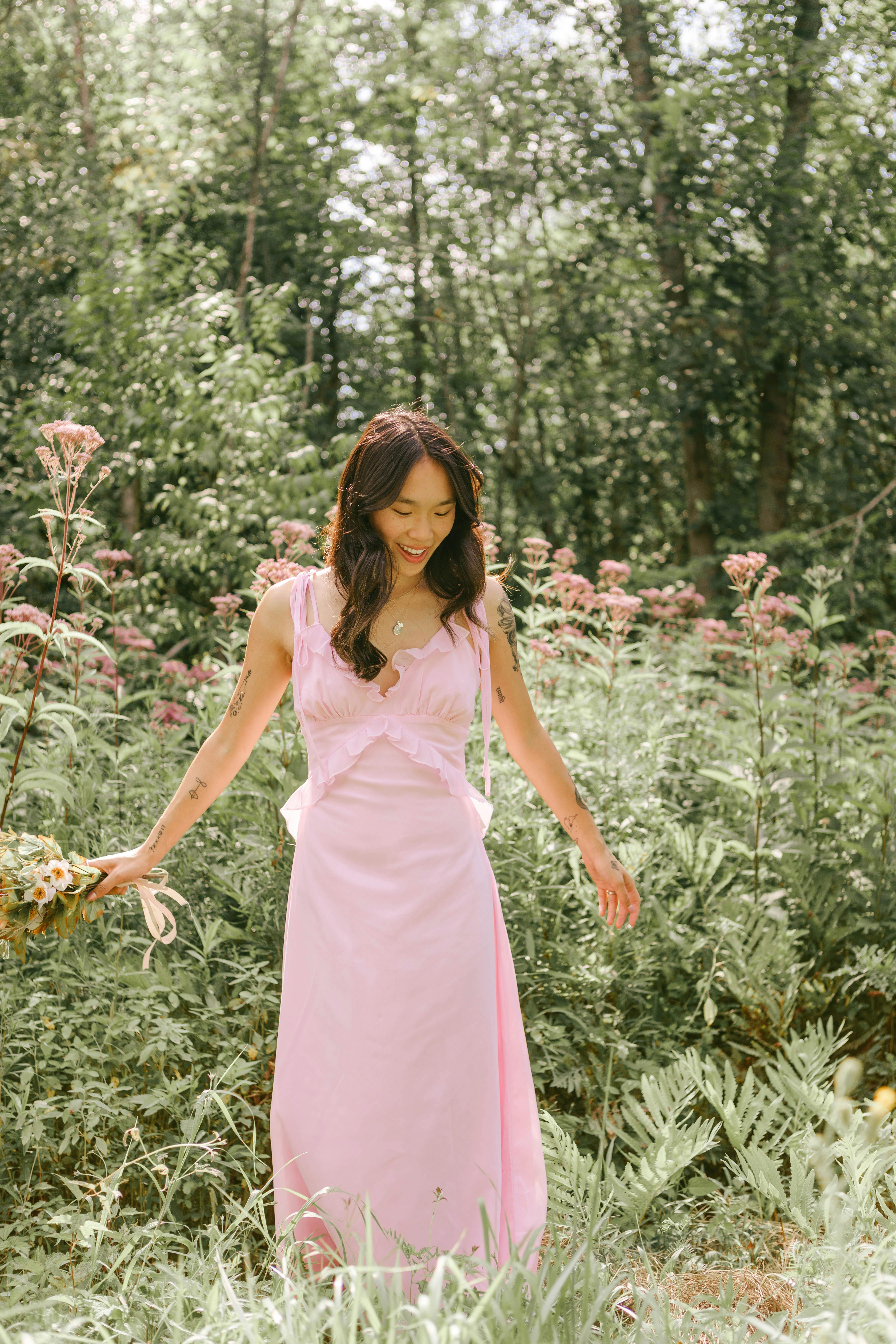 Asian woman in a pink dress smiling while walking in a lush green forest.
