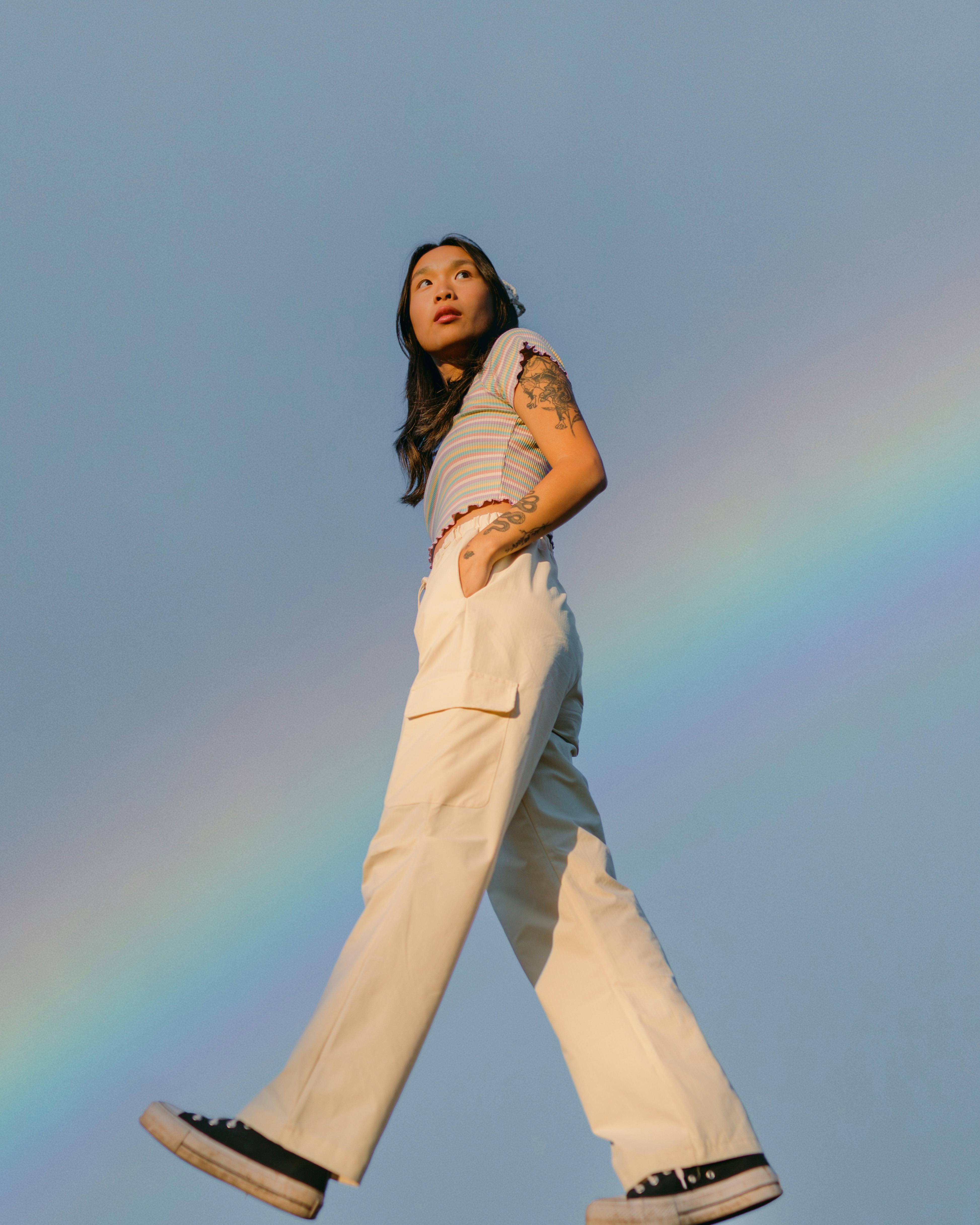 Asian woman in white pants and striped top under a vibrant rainbow in a low-angle shot.
