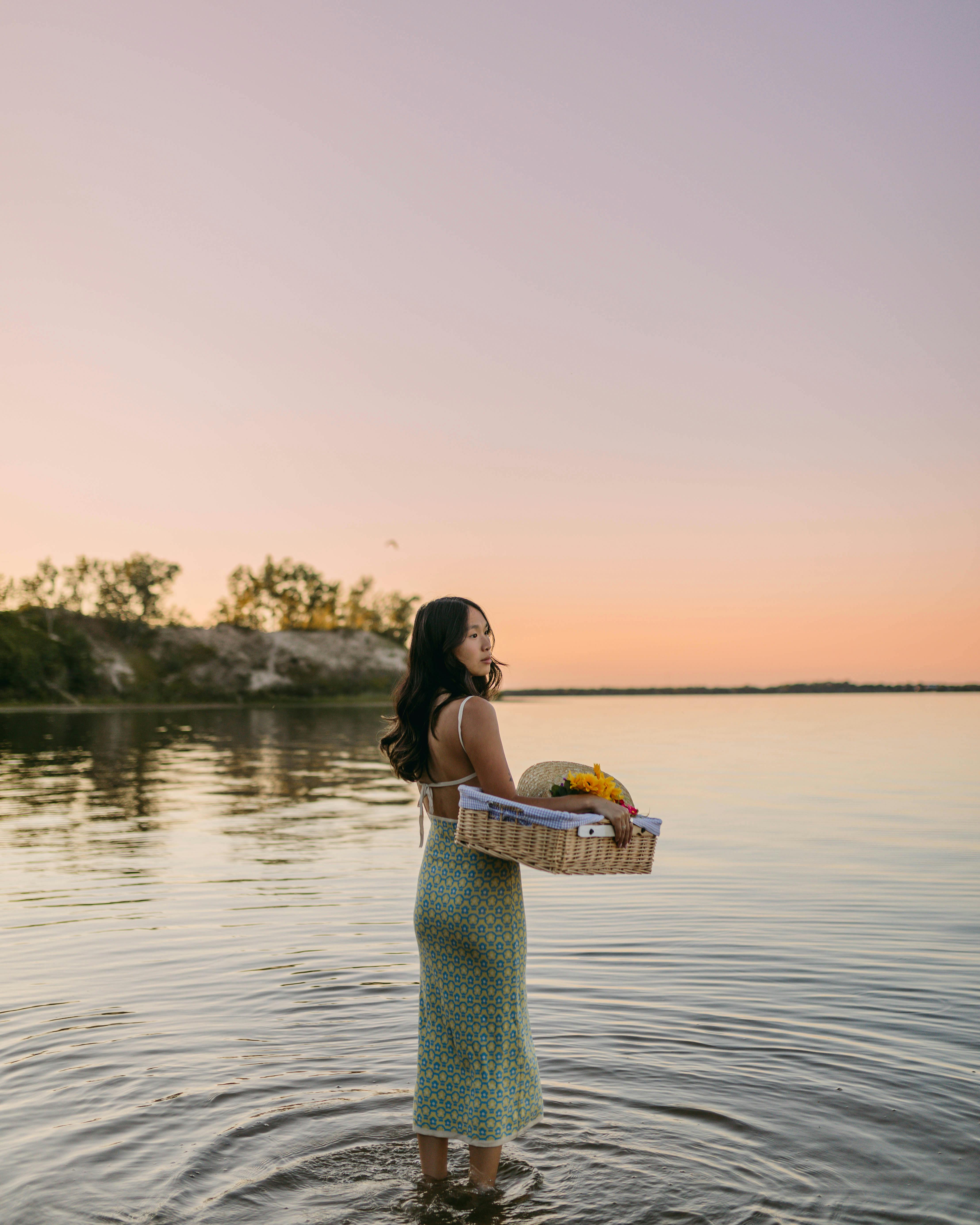 An Asian woman stands in water holding a basket of yellow flowers at sunset.