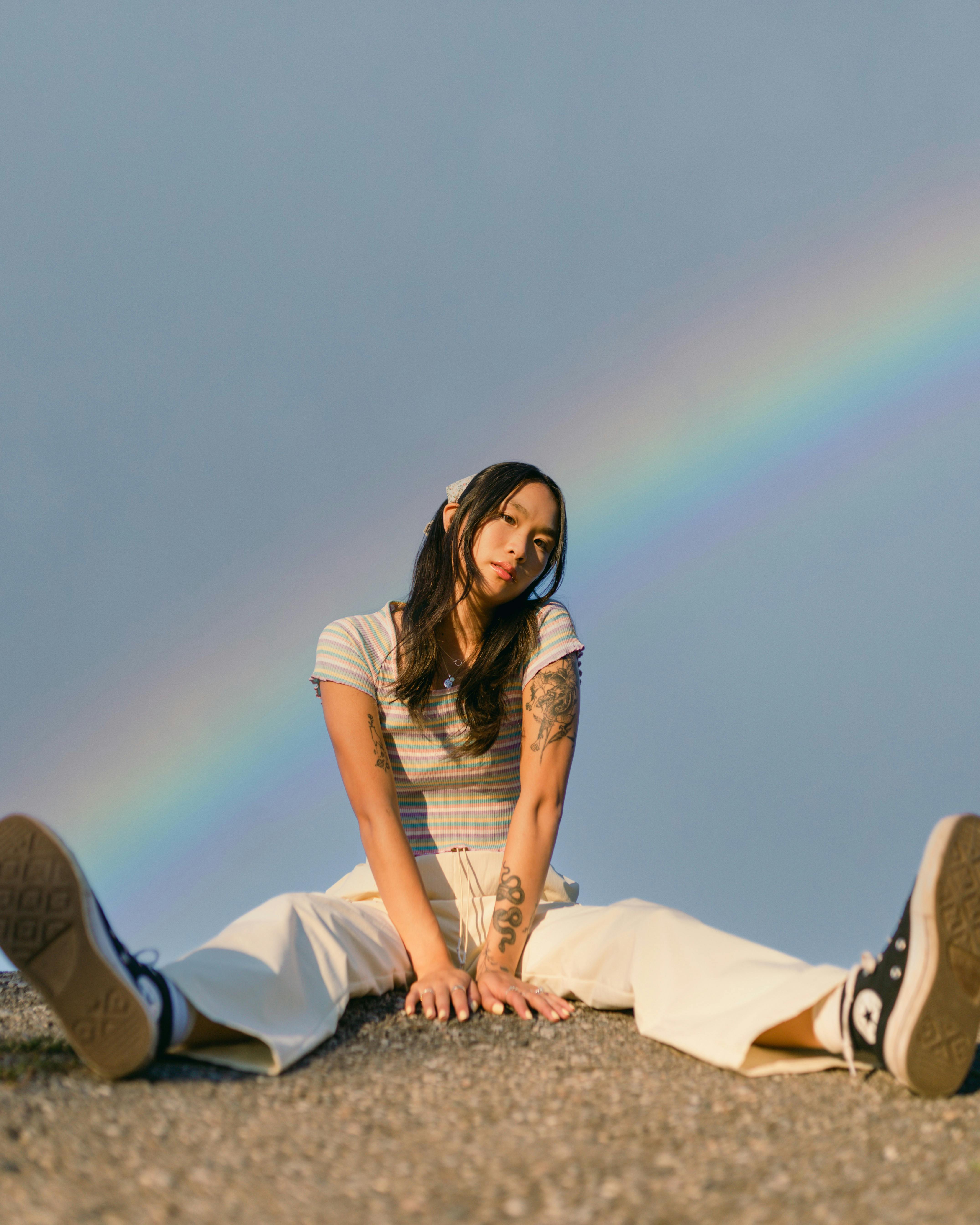 A woman sits casually outdoors with a rainbow overhead, embracing a relaxed fashion style.