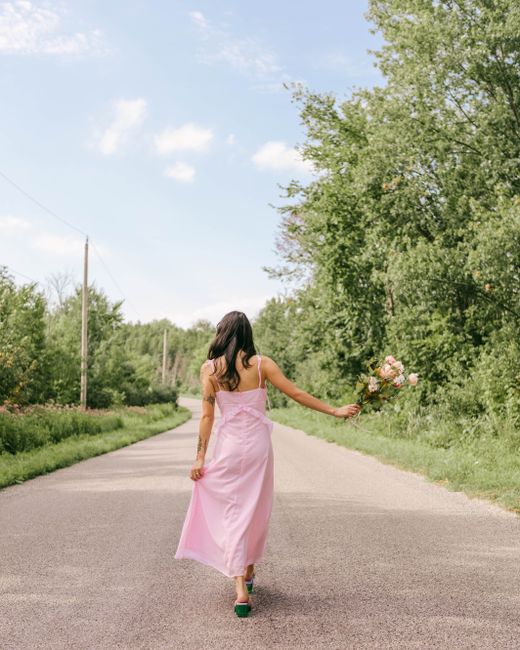 A woman in a pink dress walks on a countryside road with a bouquet of flowers.
