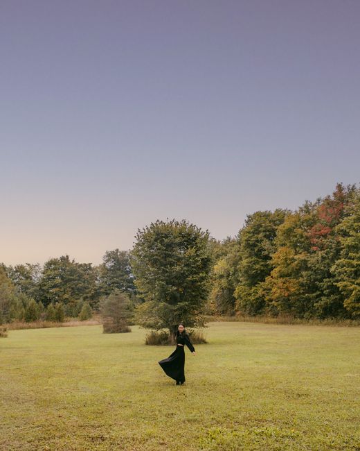A woman in a black dress is elegantly dancing in a wide-open field surrounded by trees during sunset.