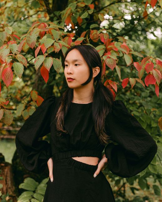 An elegant woman poses in a black dress with puff sleeves, surrounded by vibrant autumn leaves.
