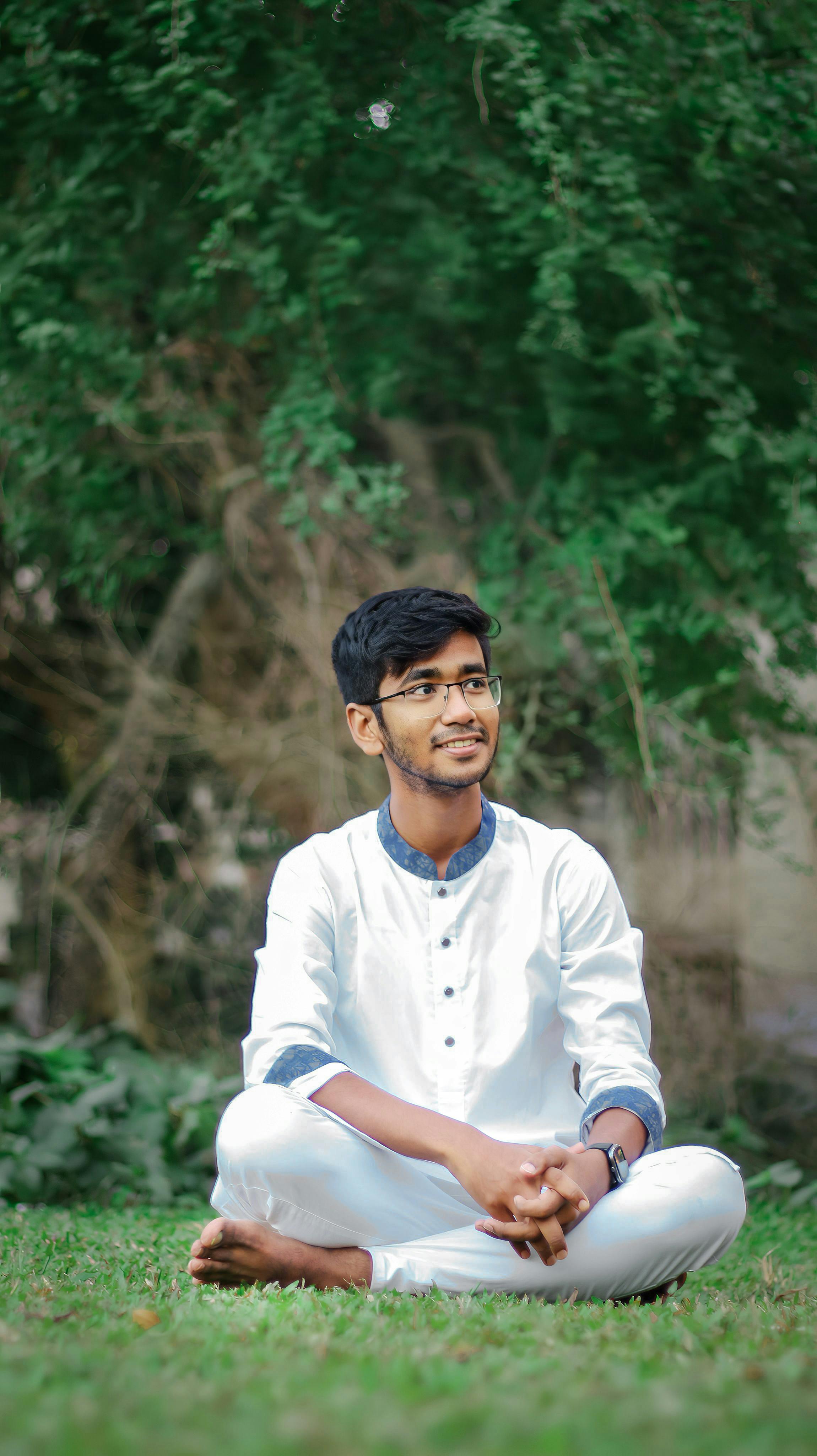 A young man in white attire smiling while sitting on grass in a serene outdoor setting.