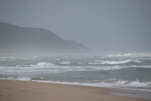 Free A serene beach scene with fog covering distant mountains and waves crashing on the shore. Stock Photo