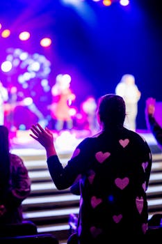 A back view of a person at a vibrant concert, with bokeh lights and performers on stage.