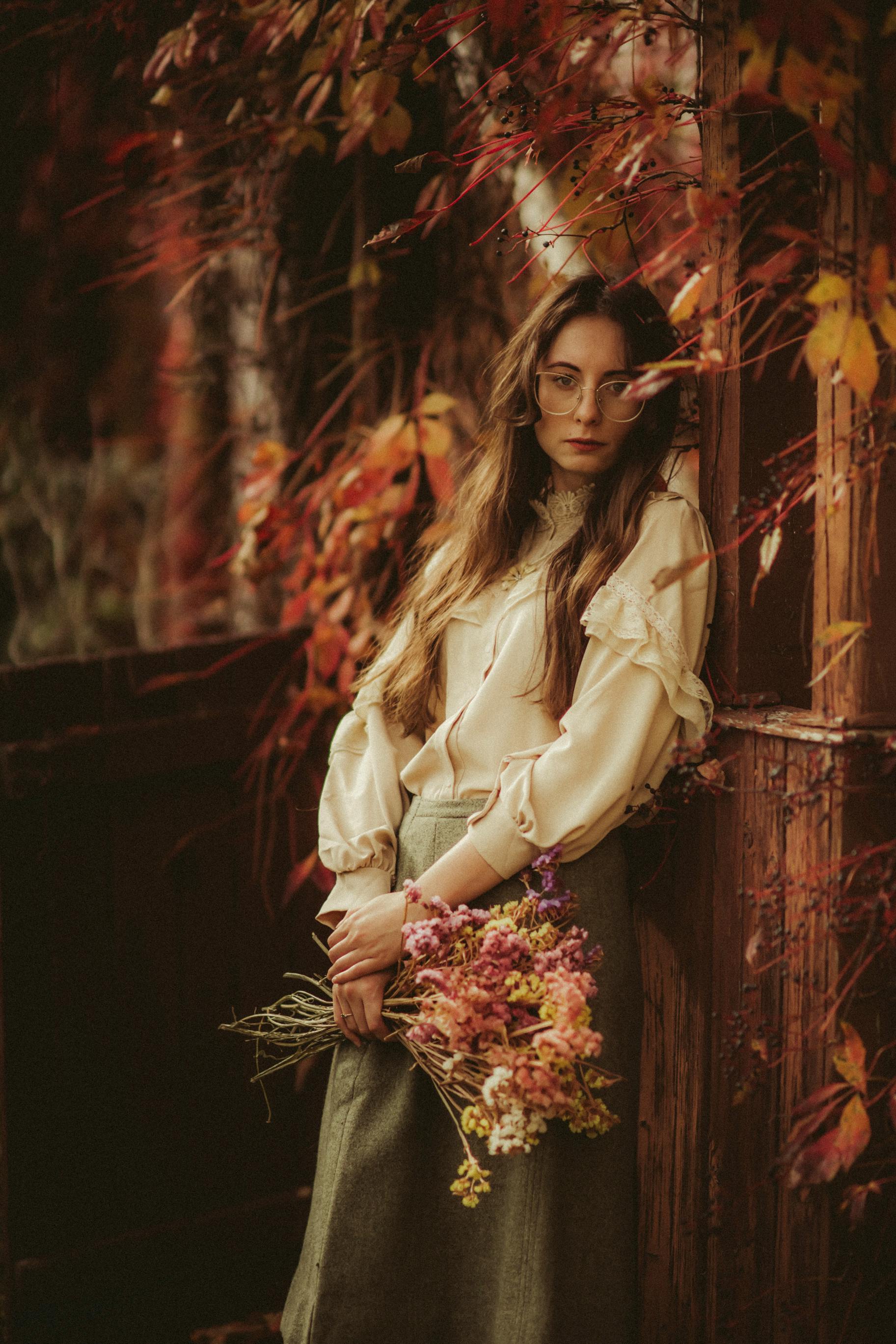 A woman in autumn attire poses gracefully with a bouquet, surrounded by vibrant fall foliage.