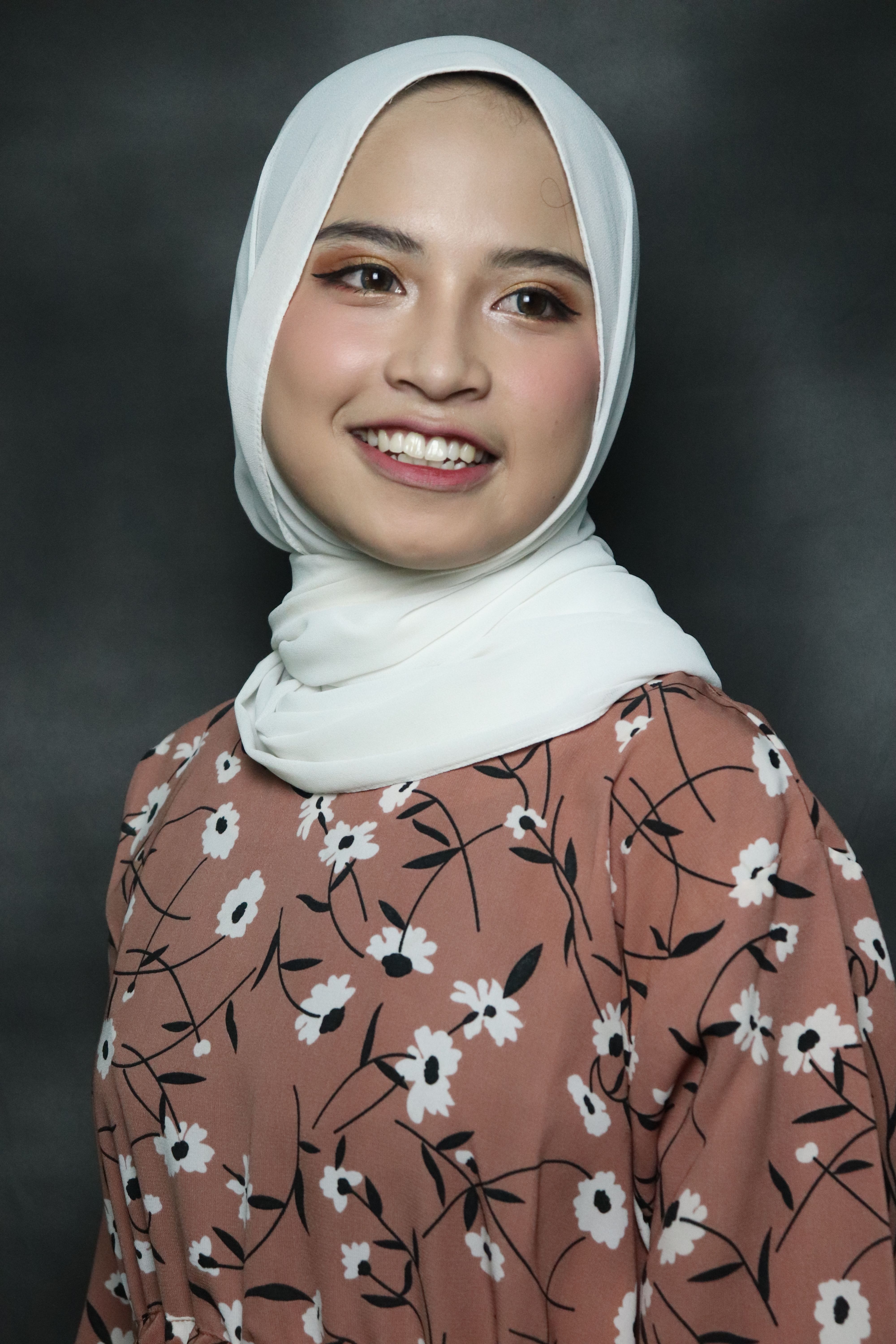 Portrait of a smiling woman wearing a floral patterned dress and hijab. Captured in studio lighting.