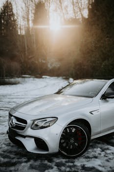 Silver Mercedes-Benz sedan parked on snow-covered ground in winter, sunlight streaming through trees.