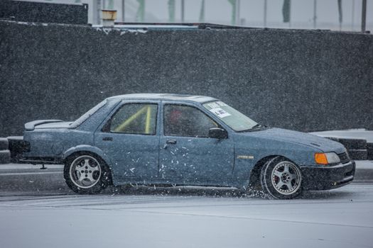 A blue car performing a drift on a snow-covered race track during winter, showcasing motorsport action.