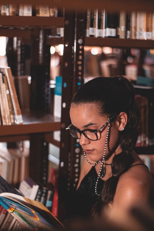 Free A young woman engrossed in a book, sitting beside bookshelves in a cozy library setting. Stock Photo