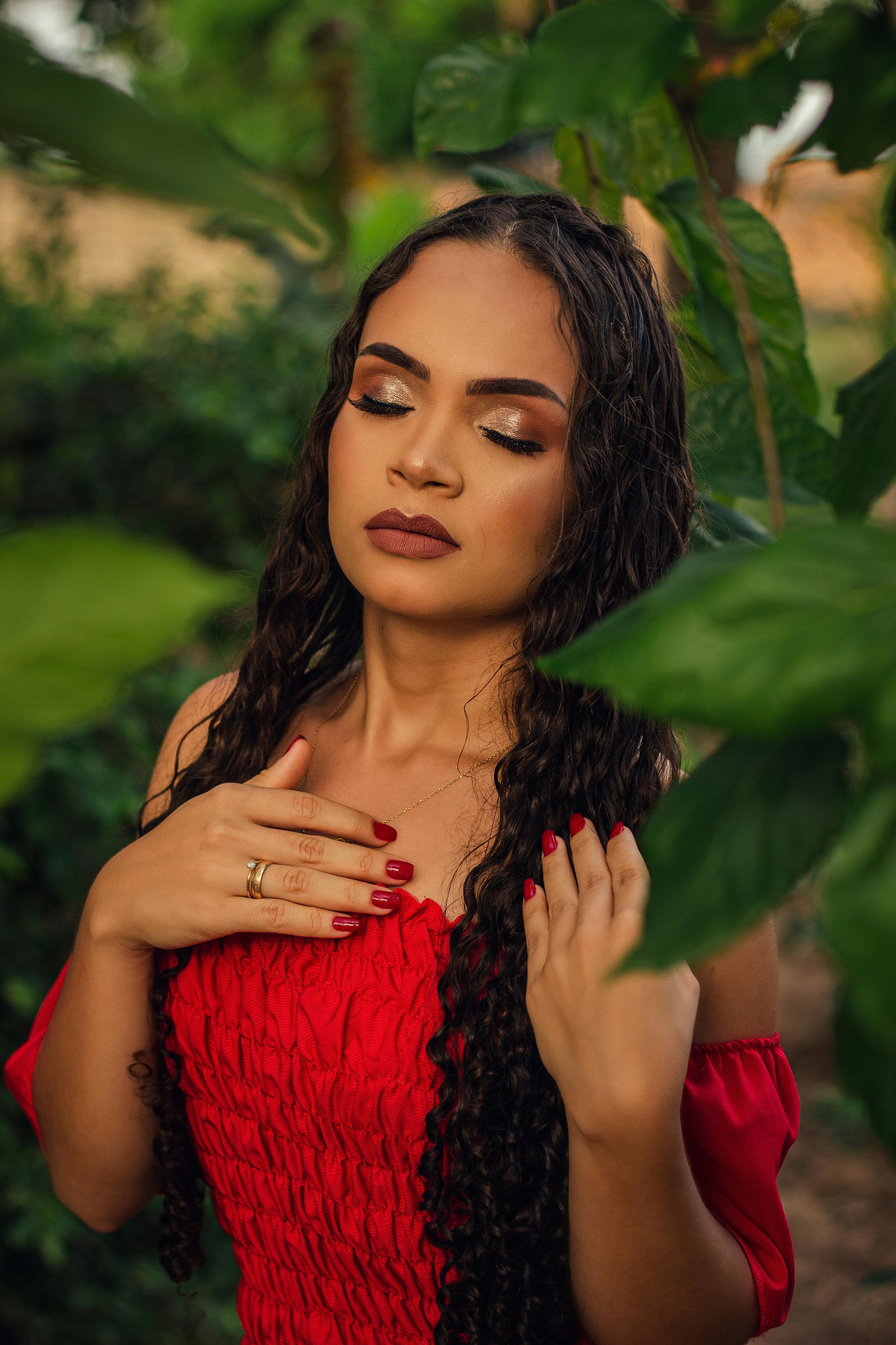 Beautiful woman with long curly hair in a red dress surrounded by greenery, eyes closed in serene expression.