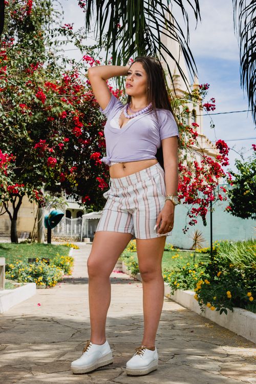 Free A fashionable woman poses confidently in a colorful garden with blooming flowers and lush trees on a sunny day. Stock Photo