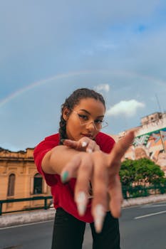 Confident woman with manicure pointing at camera under a rainbow in a vibrant urban setting.