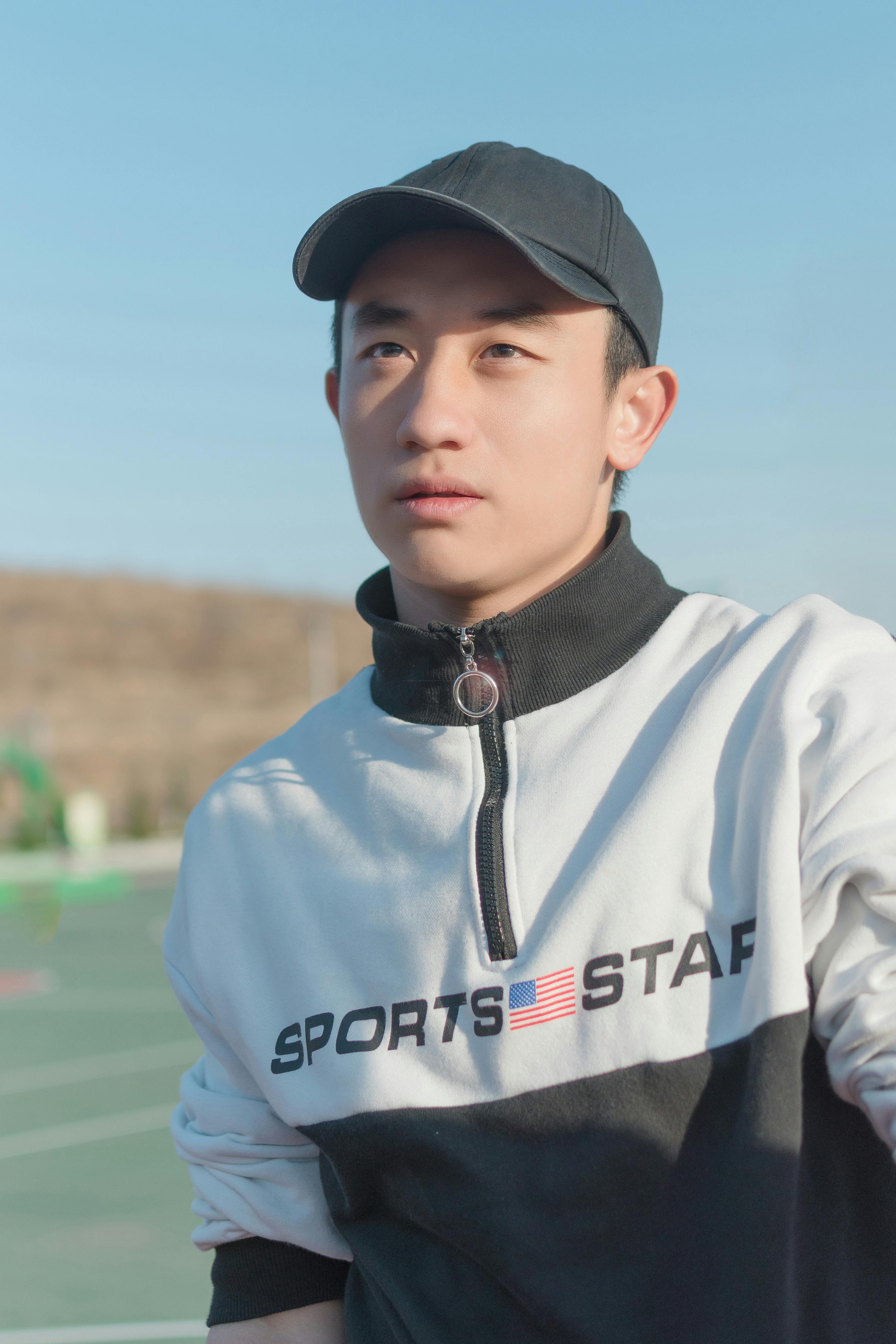 Young adult man wearing a cap and sportswear, standing outdoors under clear daylight.