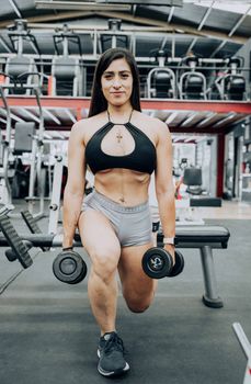 Focused woman in gymwear performing lunges with dumbbells in a modern gym.
