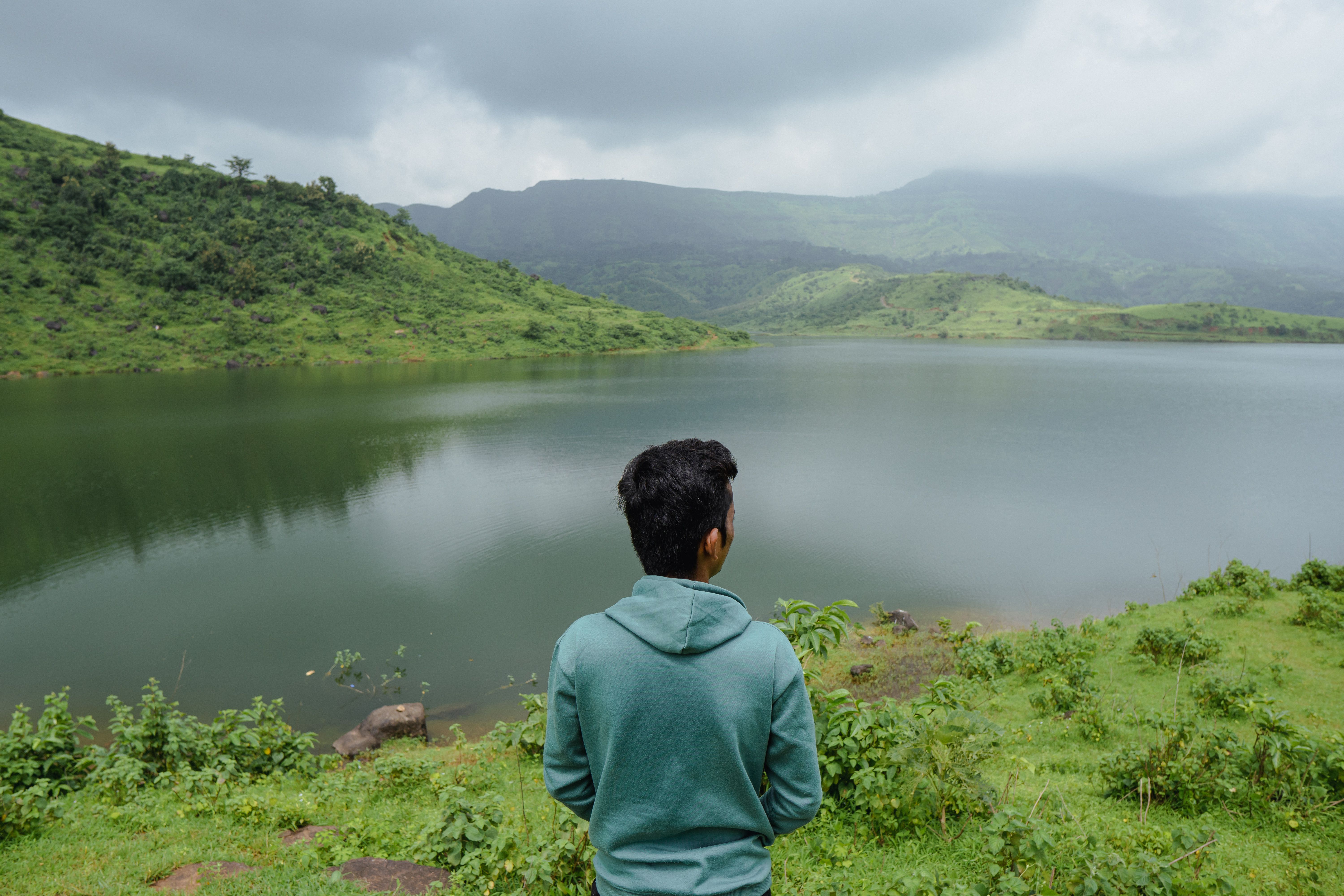 Man in hoodie observes tranquil lake and mountains in Dhom, India.