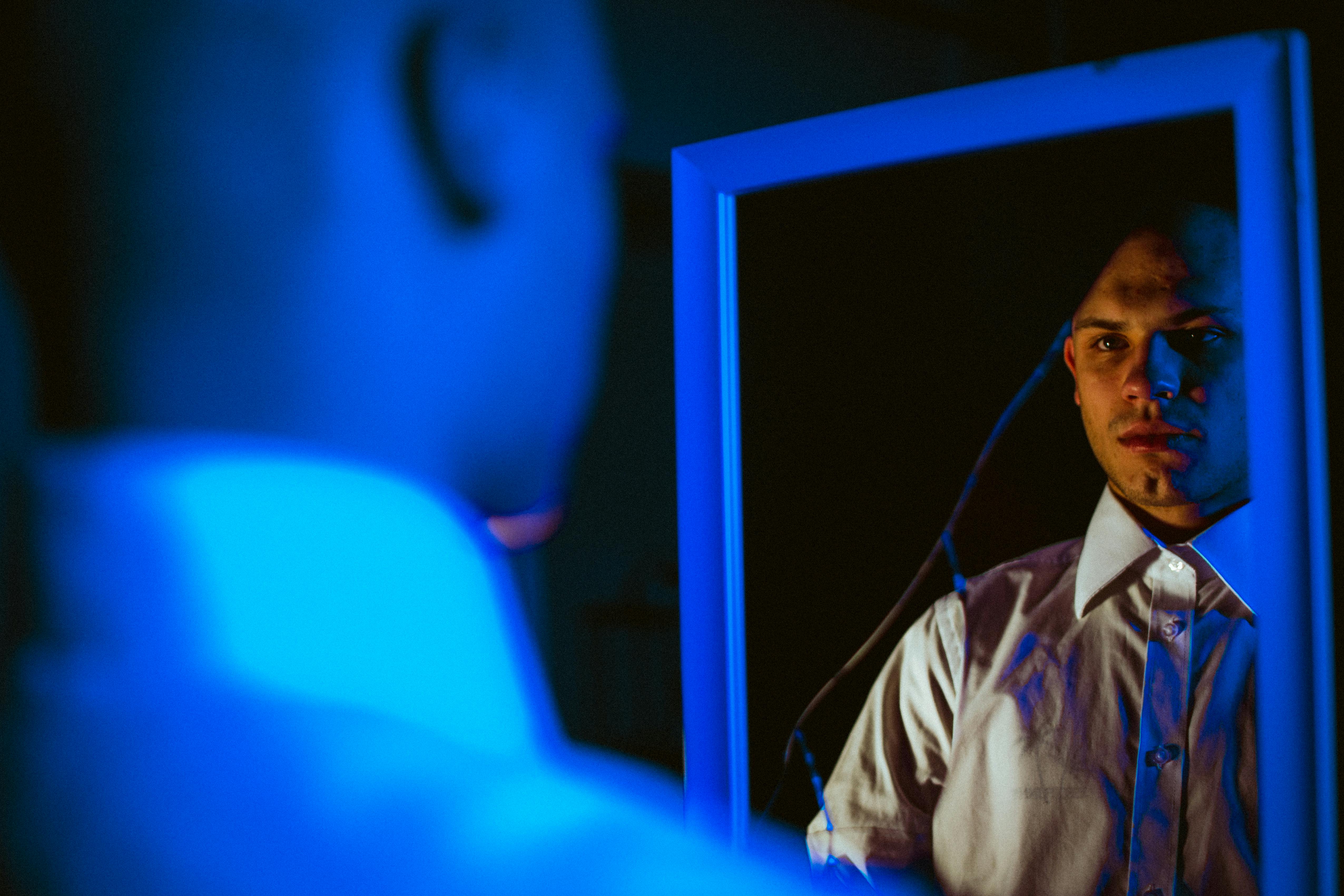A stylish man in white long sleeves looks at his reflection in a blue-lit mirror indoors.