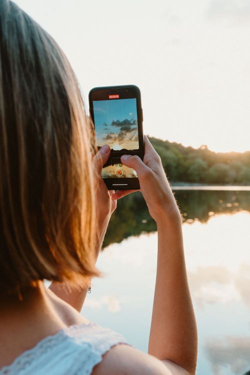 Free A woman takes a picture of a beautiful sunset over a lake using her smartphone. Stock Photo