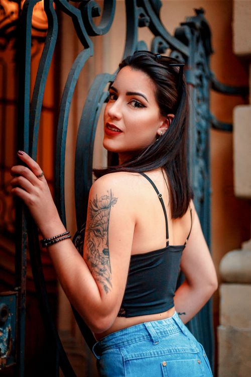 Free Elegantly posed brunette woman with tattoos by a decorative gate in an outdoor urban setting. Stock Photo