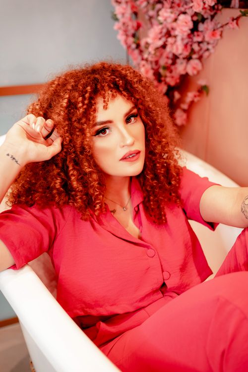 Free Chic redhead woman with curly hair wearing pink blouse, posing in bathtub with floral backdrop. Stock Photo