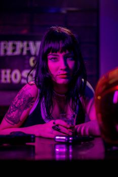 Portrait of a tattooed woman with black hair holding a drink in a neon-lit bar setting.