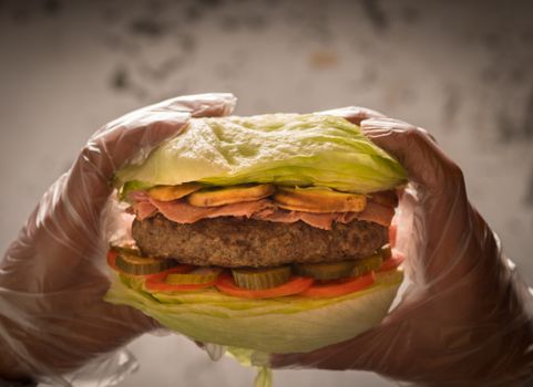 Close-up of a lettuce wrapped burger with beef patty, pickles, and tomatoes held in hands with gloves.