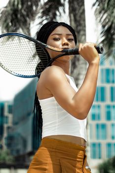 Confident African woman holding a tennis racket outdoors, showcasing fashion and sport.