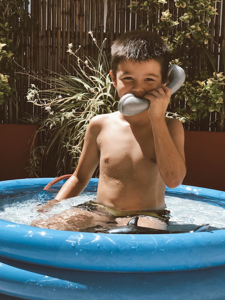 A Boy Soaking On An Inflated Pool While Holding A Telephone