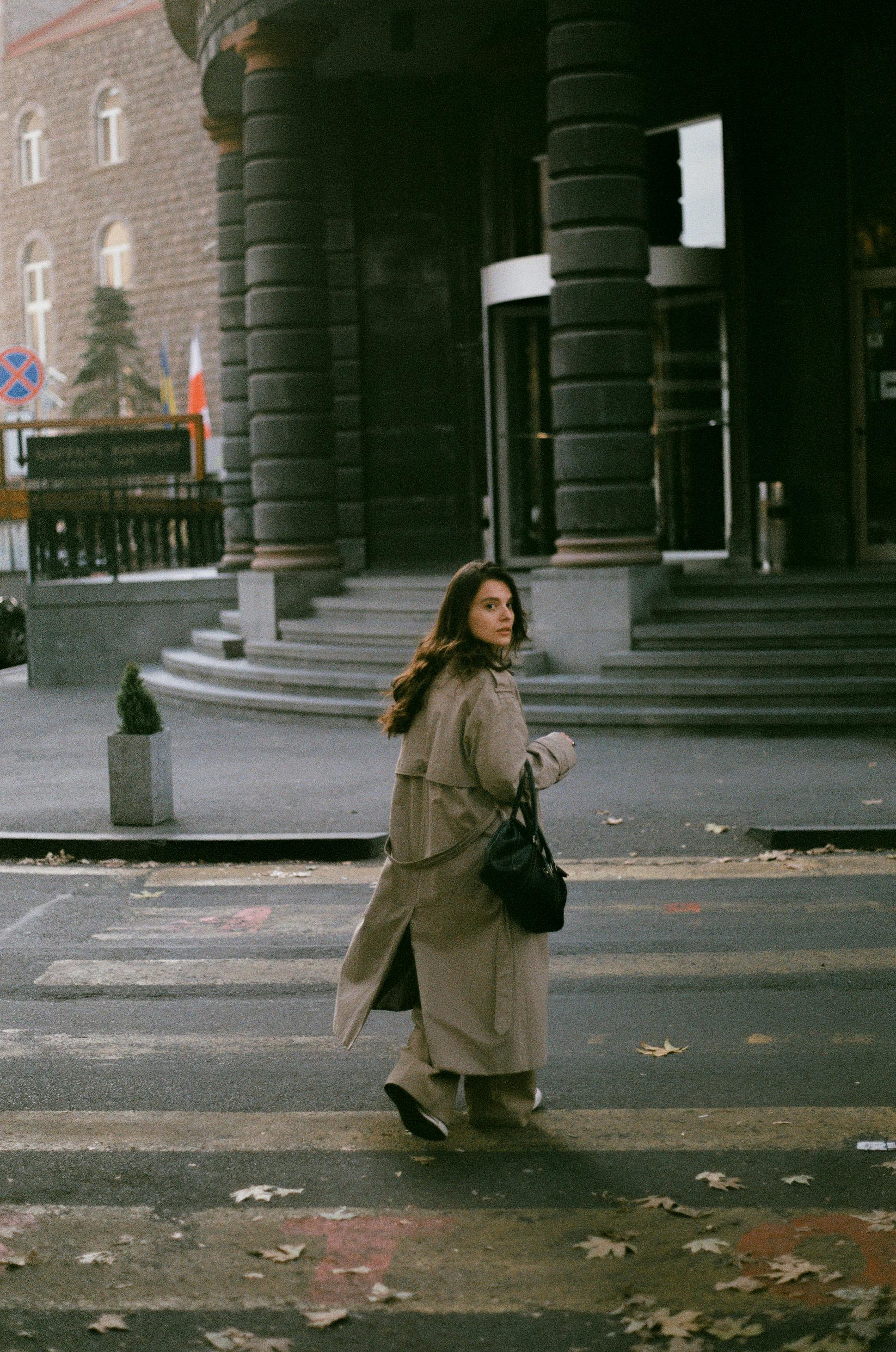 Woman in brown coat crosses city street in autumn, a cinematic urban scene captured outdoors.