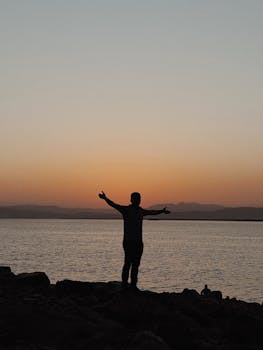 A man stands on rocky shore with arms open, silhouette against a golden sunset over water in Qurago, Iraq.