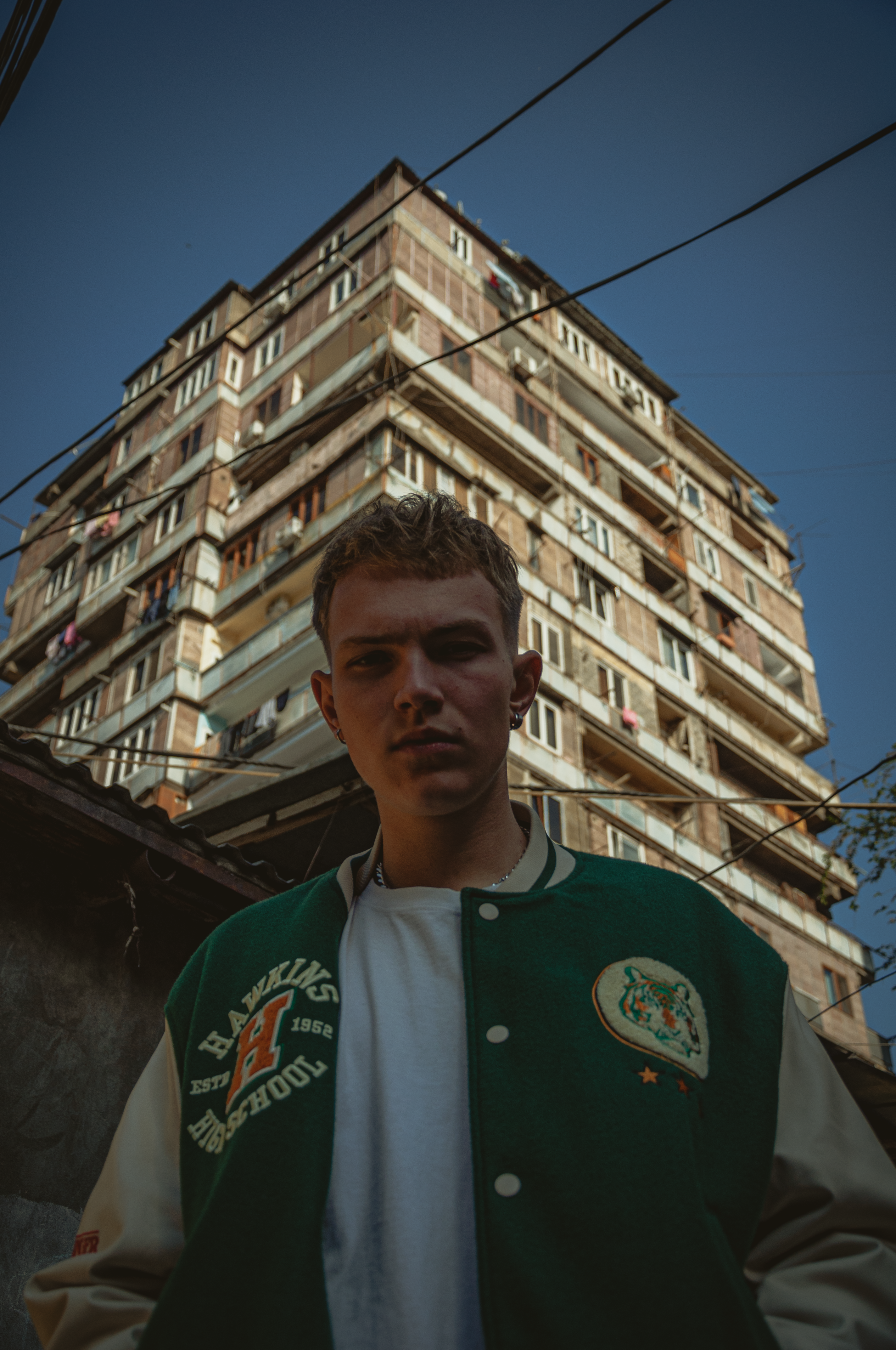 Young man in a green varsity jacket stands below a towering apartment block in Yerevan, Armenia.