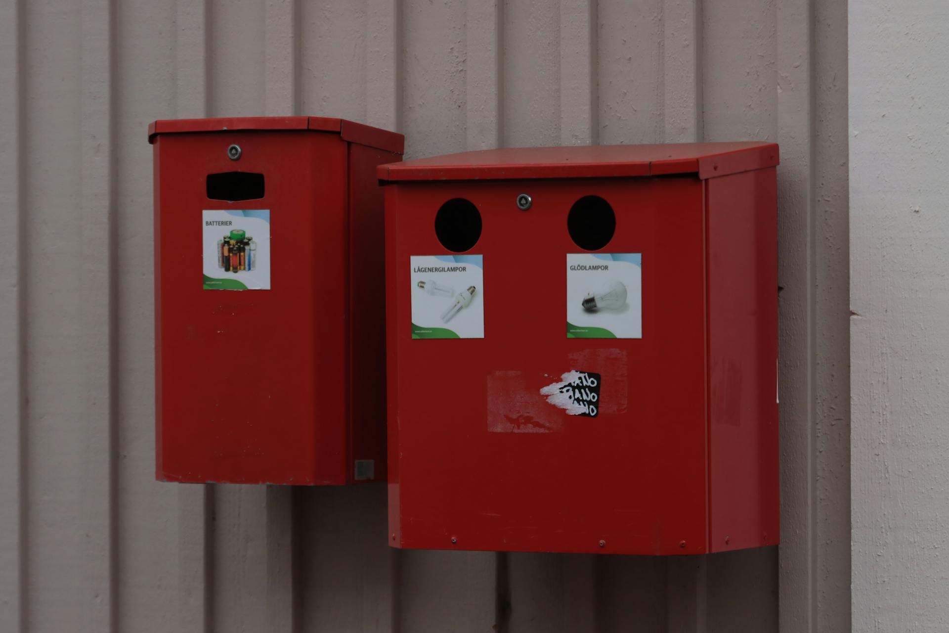 Two red recycling bins for batteries and light bulbs mounted on urban wall.