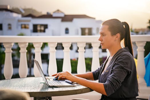 A focused woman works on her laptop outdoors on a sunny balcony, capturing a serene morning work vibe.