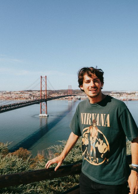Young man posing with a smile near Ponte 25 de Abril in Lisbon, under a clear blue sky.
