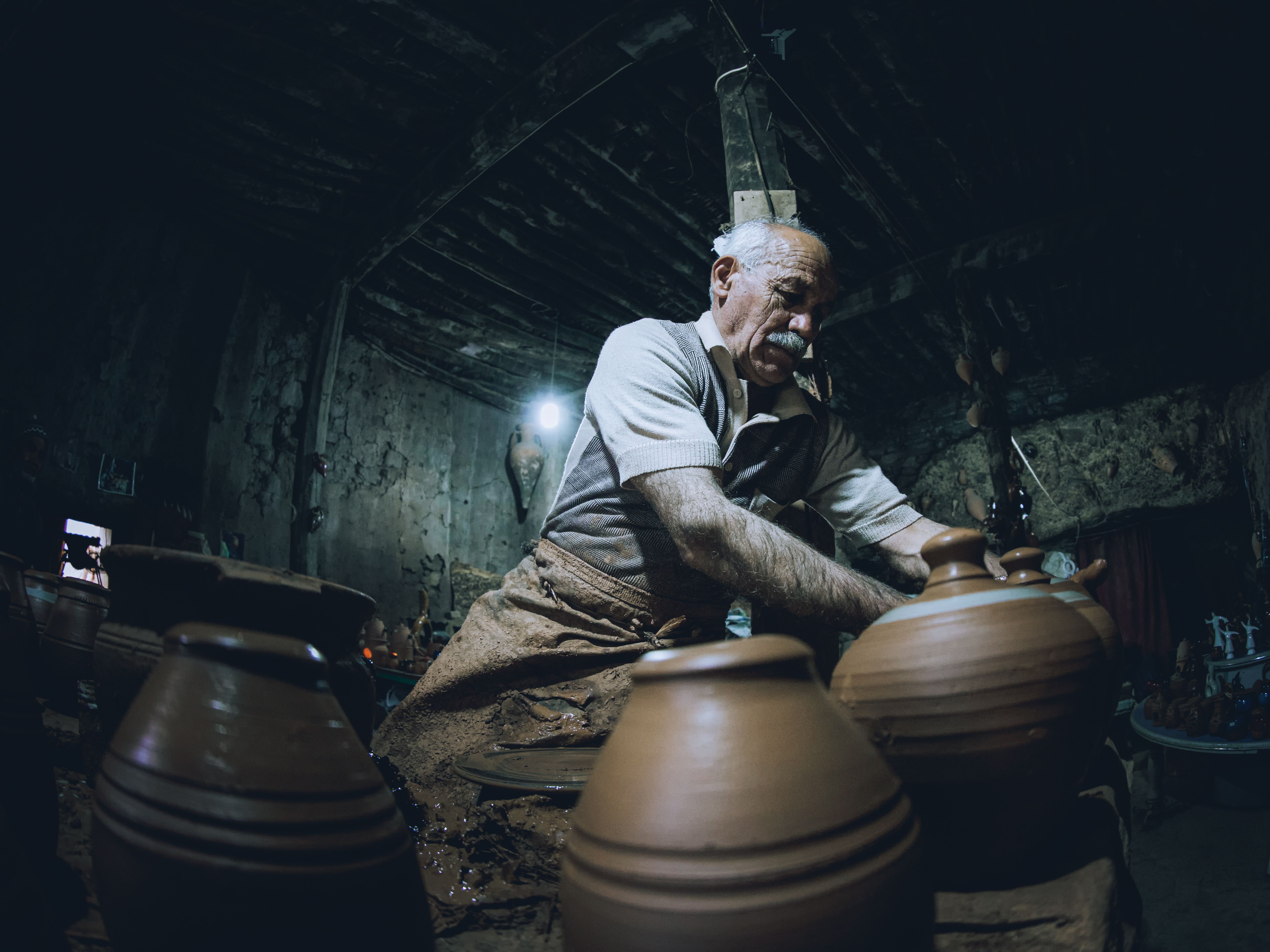 A focused elderly artisan works on pottery vases inside a dimly-lit workshop.