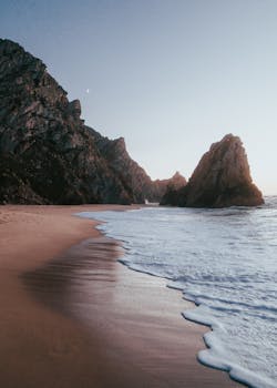 Peaceful coastal scene featuring gentle waves, rocky cliffs, and a sandy beach at sunset.