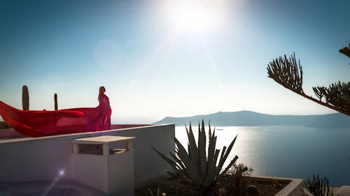 Женщина в красном длинном платье стоит на крыше здания с видом на океан