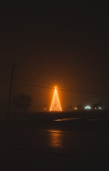 A glowing Christmas tree adorned with lights stands in the foggy night, creating a serene festive atmosphere.