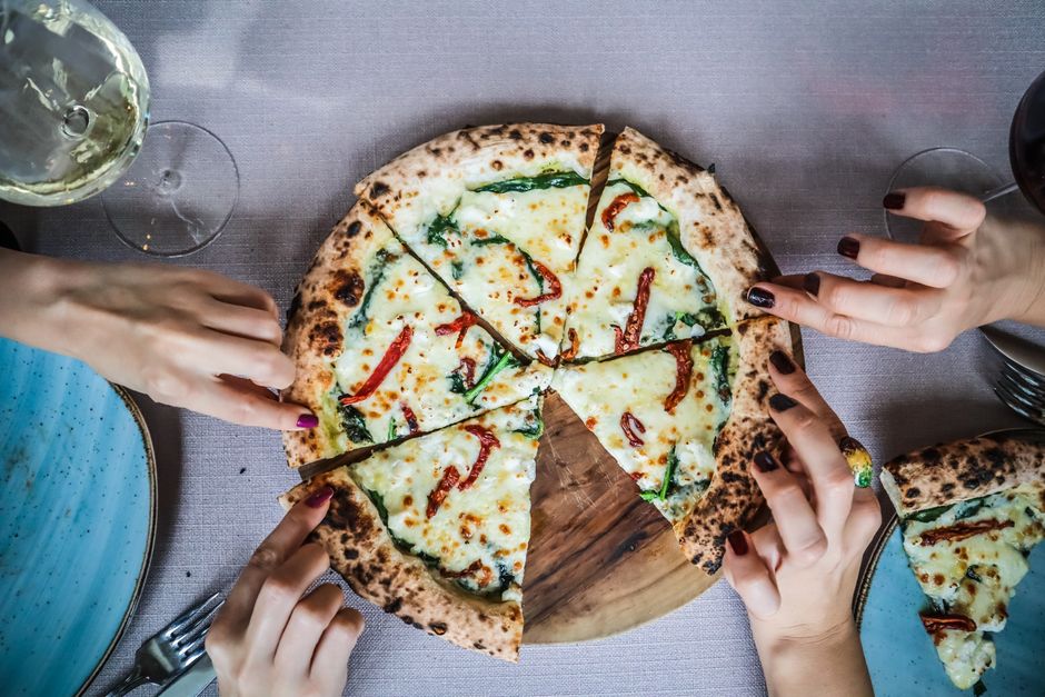 Close-up of hands grabbing gourmet pizza slices, paired with wine in a casual dining setting.