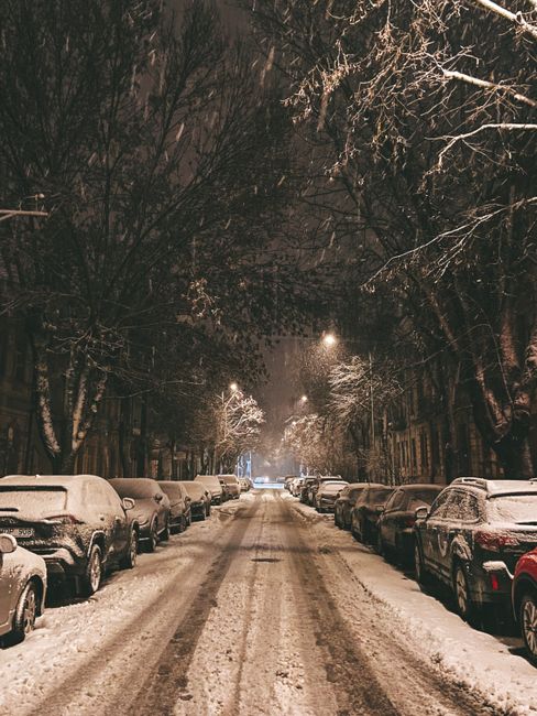 Cars parked on a snowy street at night in Vilnius, Lithuania, under streetlights.