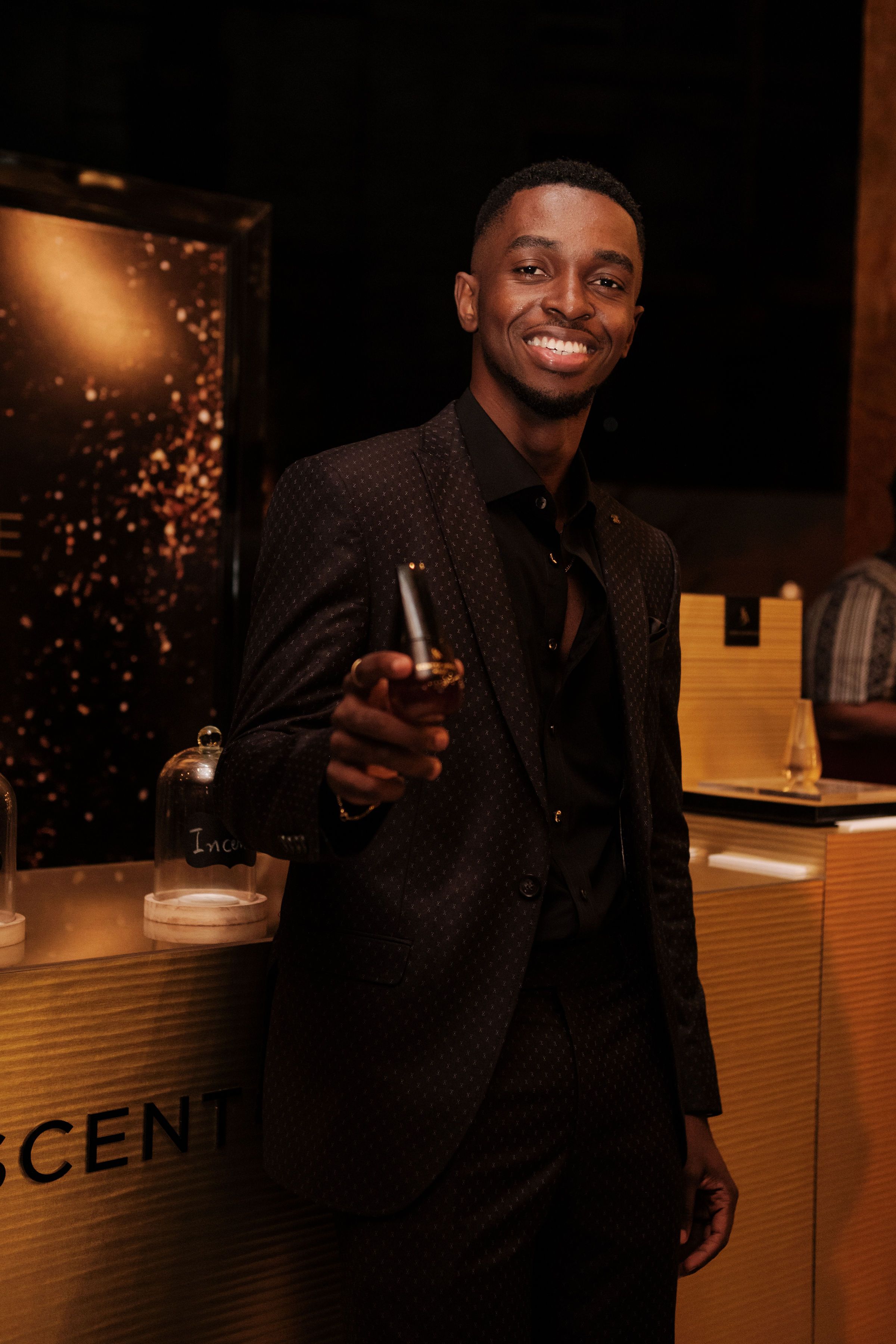 Smiling man in suit holding perfume bottle at a sophisticated indoor event.