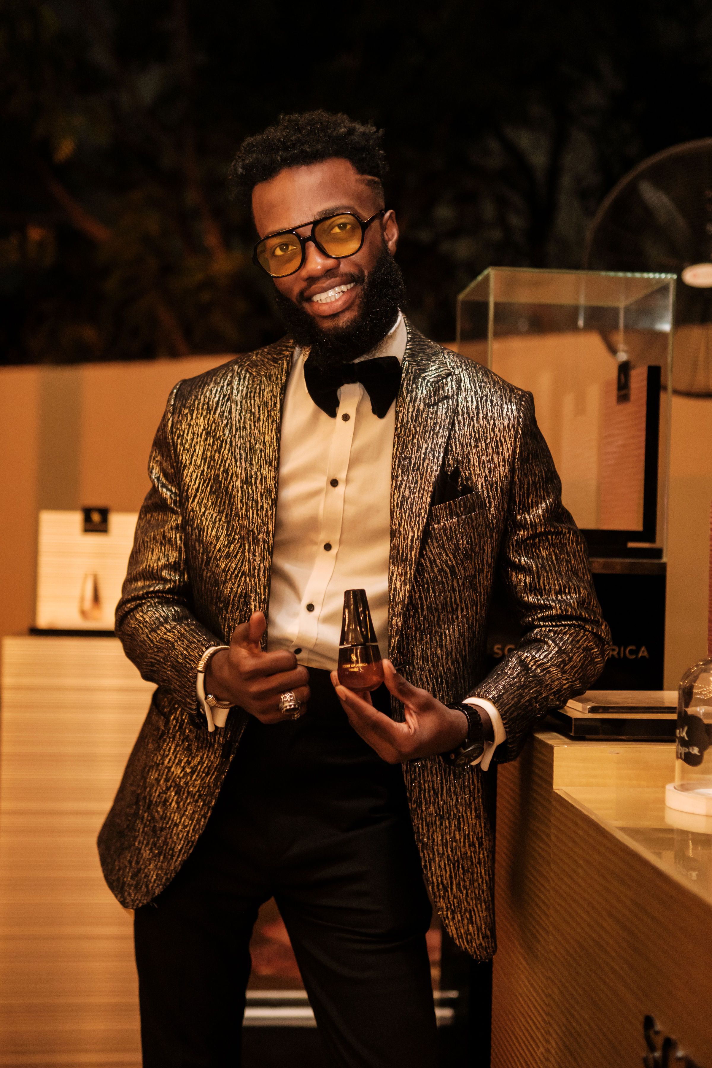 Man in a glittering suit posing with a bottle in an outdoor setting. Fashion elegance and charm.